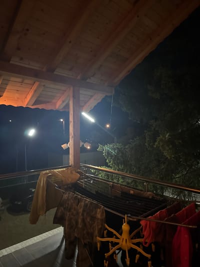 The image depicts an outdoor scene at night, likely in Ponor, Bulgaria, based on the prompt. On the left is a wooden structure, possibly a building or an awning, illuminated by warm light. Overhead, trees are visible against the dark sky. To the right, clothes are hanging on a clothesline, including a yellow plastic hanger, red fabric, tan fabric, and camouflage clothing. Surveillance cameras are mounted on a beam. The sky is dark with a few visible lights.