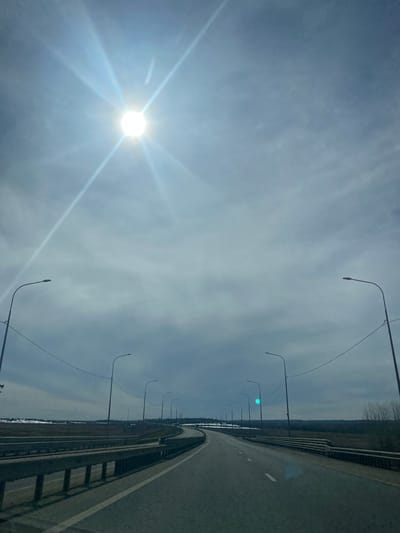 The image depicts a section of roadway in Kliuchiki, Russia. The road is gray and features white lane markings. A metal guardrail is present along the right edge of the roadway. A row of streetlights lines the left side. Beyond the guardrail on the right is a wooded area. The sky is overcast with a bright sun visible on the left.