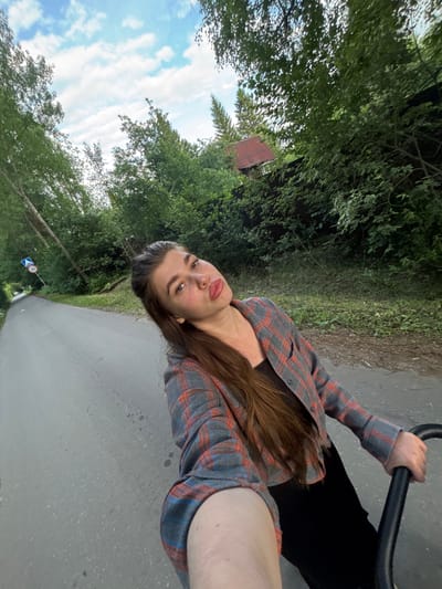 A young woman with long brown hair is taking a selfie while riding what appears to be a bicycle or scooter. She is wearing a grey and orange plaid shirt over a black top and black pants. Her left hand is on the handlebar of the vehicle. She is looking at the camera with a neutral expression. The scene is outdoors, with lush green trees lining a paved path. In the background, there is a small red structure visible among the trees, possibly a shed or a small building. The sky is blue with scattered white clouds. There is a road sign visible on the left side of the path with the number "30" and an arrow, indicating a speed limit. The image is taken from a slightly upward angle. The lighting suggests it is daytime, likely late afternoon or early evening, with soft light filtering through the trees. The overall atmosphere is casual and relaxed, suggesting a leisurely ride in a park or rural area. The location context is Шевлягино, Russia.