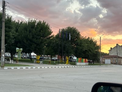 The image captures a street scene in the city of Letnitsa, Bulgaria, during twilight. The sky is filled with dramatic, colorful clouds, transitioning from pinks and oranges to grays. In the foreground, a paved road curves to the right, with a pedestrian crossing marked by white and gray stripes. On the left side of the road, a concrete utility pole with overhead wires stands tall. Adjacent to the road is a grassy area with benches. One bench, on the far left, is adorned with a large, light green bow. Another bench, further to the right, is decorated with yellow sunflower motifs. Behind the benches, a line of lush green trees forms a dense backdrop. Several cars are parked along the street behind the trees. In the center of the image, three flagpoles stand, with flags of Italy, Bulgaria, and the European Union visible. Further to the right, a colorful sign spells out "ЛЕТНИЦА" (Letnitsa) in large, vibrant letters. Beyond the sign, a cluster of yellow sunflower figures is present. To the far right, a brick building with a tiled roof and a large grey door is visible, along with a streetlamp. The overall impression is a town square or a main street in a Bulgarian town as daylight fades.