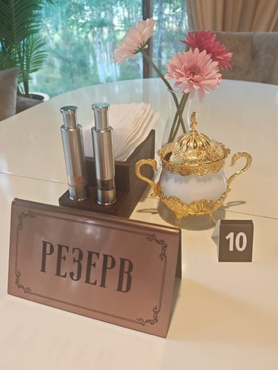This image shows a table setting in what appears to be a restaurant. The table is white and set with several items. In the foreground, a rose-gold colored sign reads "РЕЗЕРВ" (RESERVE) in a decorative font, indicating that the table is reserved. To the right of the sign, a small brown tent card displays the number "10" in white, likely a table number.
Further back on the table, there's a decorative gold and white sugar bowl with a lid, featuring ornate carvings and handles, sitting on a white surface. Next to it, a dark wooden holder contains a stack of white napkins and two silver salt and pepper shakers. Behind these items, a clear glass vase holds three artificial flowers: two pink and one lighter pink.
The background suggests an indoor setting with a large window or glass wall offering a view of greenery and trees, possibly indicating a location on the outskirts of a city or in a natural setting. The lighting is soft, suggesting either natural light from the window or ambient indoor lighting. The overall impression is one of a clean, well-maintained dining space. The location is described as being in the city of Novy Urengoy, Russia.