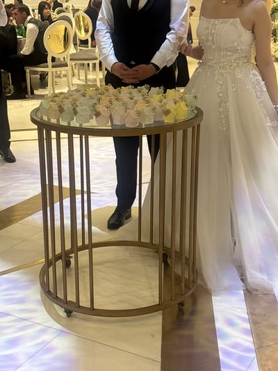 This appears to be a wedding reception scene, possibly indoors, in a location that may be in the city of Masis, Armenia. A bride in a white wedding dress is standing next to a groom wearing a dark suit and white shirt with a bow tie. They are both standing near a round table with a mirror top. The table is surrounded by a metal frame with decorative detailing and is holding small, decorated boxes or favors. The lighting is bright, with a pattern of what appears to be colored light projections, possibly from stage lighting, on the floor. Around the table, other people can be seen seated on chairs.
