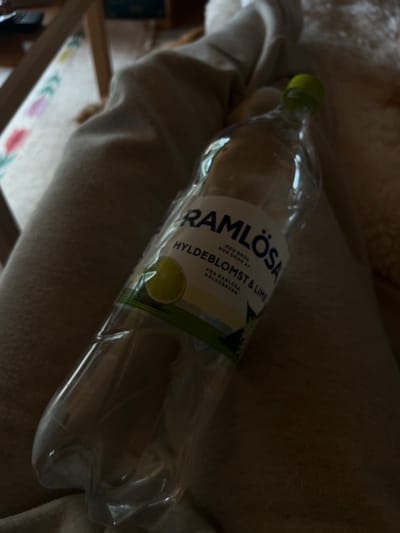 The image shows a glass bottle of Ramlösa Hyldeblomst & Lime soda resting on a pair of tan pants. The bottle is laying on its side, with the label facing up. The label has text that is partially obscured but can be read as "Ramlösa med brus med smak av Hyldeblomst & Lime". It also says "Från Ramlösa Hällsokbrunn". There is an image of a lime on the label. The bottle is partially filled with clear liquid. The background is a light brown color. The scene is located in the city of Blåvand, Denmark, at the coordinates 55.558, 8.158.