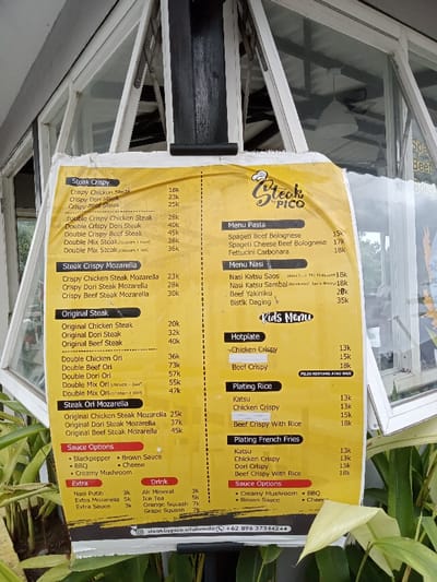 A yellow, worn menu board for "Steak by PICO" is prominently displayed, likely outdoors at a restaurant or cafe in Kecamatan Panji, Indonesia. The menu is printed on a bright yellow background with black text and some red accents for headings like "Sauce Options" and "Kids Menu." It's attached to a black vertical pole, with a multi-paneled glass window structure visible behind it, and lush green plants at its base.
The menu is divided into several categories:
* **Steak Crispy** and **Steak Crispy Mozarella** sections list items like Crispy Chicken Steak (18k), Crispy Dori Steak (23k), Crispy Beef Steak (25k), and double portions or mozzarella-added versions with varying prices up to 45k.
* **Original Steak** and **Steak Ori Mozarella** feature Original Chicken Steak (20k), Original Dori Steak (32k), and Original Beef Steak (40k), also with double and mozzarella options, reaching up to 73k for Double Beef Ori.
* **Menu Pasta** includes Spageti Beef Bolognese (15k), Spageti Cheese Beef Bolognese (17k), and Fettucini Carbonara (18k).
* **Menu Nasi** (Rice Menu) offers Nasi Katsu Saos (18k), Nasi Katsu Sambal (18k), Beef Yakiniku (28k), and Bistik Daging (35k).
* A **Kids Menu** section presents Hotplate items like Chicken Crispy (13k), an unnamed 15k item, and Beef Crispy (18k), noting "PILIH KENTANG ATAU NASI" (Choose Fries or Rice).
* **Plating Rice** and **Plating French Fries** offer Katsu (13k), Chicken Crispy (13k), Dori Crispy (15k), and Beef Crispy With Rice (18k).
* **Sauce Options** include Blackpepper, Brown Sauce, BBQ, Cheese, and Creamy Mushroom.
* **Extra** items are Nasi Putih (3k), Extra Mozarella (5k), and Extra Sauce (3k).
* **Drinks** available are Air Mineral (3k), Ice Tea (5k), Orange Squash (7k), and Grape Squash (7k).
The prices are denoted with 'k', indicating thousands of Indonesian Rupiah. Contact information, "@steakbypico.situbondo" and phone number "+62 896 37344244", is visible at the bottom. The overall scene suggests a casual dining establishment displaying its offerings during daylight hours.
