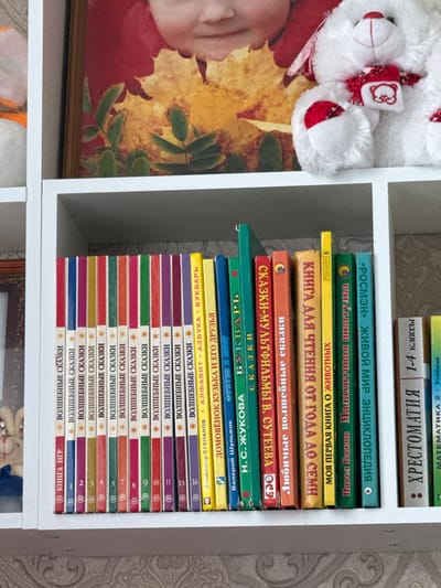 The image shows a white shelf filled with books. The books are of various colors and sizes, and they are arranged in a row. Some of the books have titles in Russian. In the background, there is a framed picture of a child with autumn leaves, and a white stuffed animal with a red scarf. The shelf is in a room with a textured wallpaper. The overall scene depicts a home library or a collection of children's books. The text on the books and the context suggest this location is in Rossosh, Russia.
