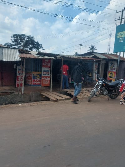 The image depicts a street scene in Kisanga, Tanzania. An asphalt road occupies the foreground, leading to a row of informal, single-story shops or stalls with corrugated metal roofs. A concrete drainage channel separates the road from the structures, bridged by three wooden planks allowing access. The shopfronts are characterized by multiple advertisements for mobile money services, including Airtel, NMB WAKALA, M-pesa, Moode, and Halo Pesa.
Three individuals are present. A male subject, seen from behind, wears a dark jacket, light blue jeans, and black sandals, standing near the road's edge, seemingly positioned to enter the shops. Inside one of the open stalls, a second male subject, wearing a red t-shirt and blue trousers, is partially visible with his back mostly to the camera. On the far right, the lower body of a third person, dressed in patterned fabric, is partially obscured by a black motorcycle. This motorcycle is parked facing left, positioned at the road's edge directly in front of the shops. Overhead, numerous utility lines traverse an overcast sky with patches of blue. In the background, a blue billboard with white text, partially legible as "OKTOBE TUNATI...", is visible on the right, alongside various trees.