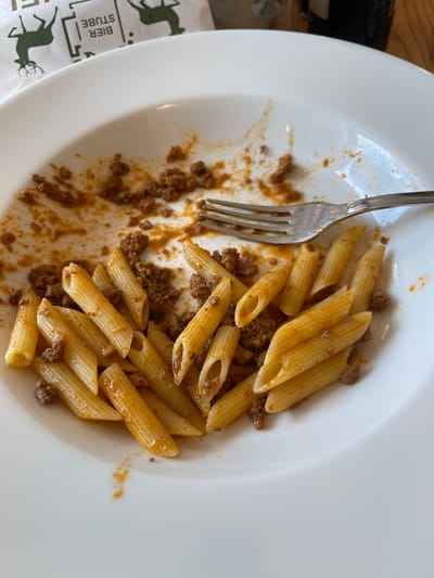 A white plate of penne pasta with a meat sauce is shown from a top-down perspective. The pasta is cooked al dente and coated in a rich, reddish-brown sauce with crumbled meat. A silver fork is positioned on the left side of the plate, partially immersed in the pasta and sauce. The plate is mostly full, with some sauce and meat scattered around the edges, suggesting the meal is in progress or has recently been enjoyed.
To the left of the plate, a white paper placemat with a dark green graphic is visible. The graphic features two stylized deer silhouettes and text that reads "BIER STUBE". This indicates a possible setting in a brewery or beer hall. The overall lighting suggests daytime. The image is likely taken indoors, possibly in a restaurant or dining area in Cortina d'Ampezzo, Italy. There are no people visible in the image.