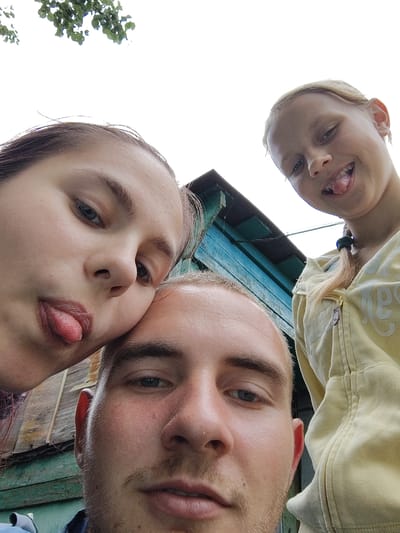 A low-angle selfie captures three individuals in what appears to be a rural or outdoor setting, possibly near a weathered wooden structure and a teal-colored outbuilding. In the foreground, a man's face is prominently featured, with his eyes looking upwards and slightly to the left. To his left, a young woman's face is angled towards the camera, her eyes looking to the side with her tongue playfully sticking out. To the right and slightly behind the man, a younger girl with blonde hair tied in a ponytail grins at the camera, also with her tongue sticking out. She is wearing a light yellow hooded jacket. The background reveals a mix of wooden planks and the painted exterior of the teal building, suggesting an outbuilding or shed. The sky is visible in the upper portion of the image, with some green leaves from a tree peeking into the frame from the top left. The overall impression is a casual, playful moment captured in an outdoor environment. The mention of Okhtyrka, Ukraine, suggests a possible geographical context for this scene.