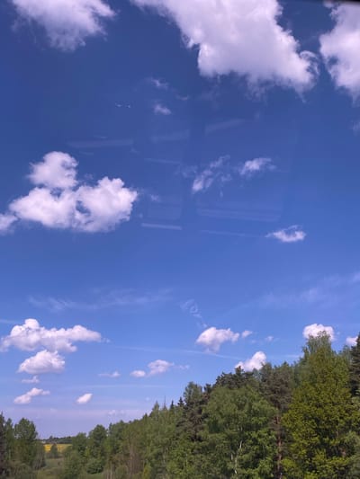 The image shows a view of the sky with scattered white cumulus clouds and a treeline in the right third of the frame. The sky is a vibrant blue. There are reflections on the left side of the image, possibly from a window. Based on visual cues, the image may have been captured from a vehicle. There are no discernible people, actions, or interactions within the frame. The geographical location is Лешково, Russia, inferred from available data. The environment consists of a blue sky, clouds, and trees.