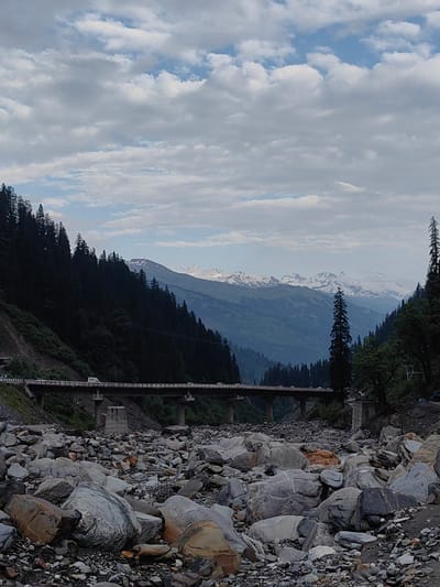 The image captures a wide-angle view of a mountainous landscape in Dhundi, India. In the foreground, a dry, rocky riverbed is filled with a multitude of grey and brown stones of various sizes. Beyond the riverbed, a long, elevated concrete bridge spans across the terrain, with several vehicles visible on its surface. The bridge is supported by concrete pillars. To the left of the bridge, a steep, dark green forested mountainside rises sharply, covered in dense coniferous trees. Further in the distance, more layers of mountains are visible, with the peaks of some mountains capped with snow. The sky above is partly cloudy, with patches of blue peeking through. The overall scene depicts a remote, natural environment with human infrastructure integrated into the landscape.