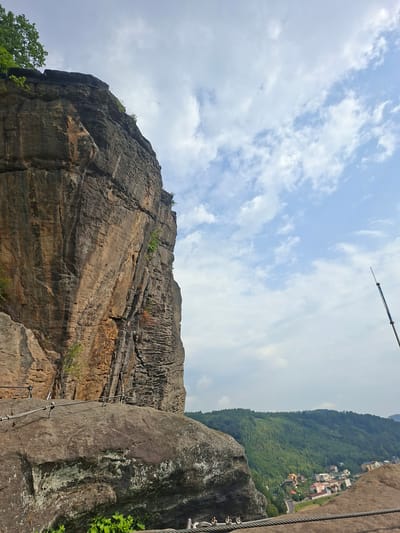 The image depicts a dramatic cliff face on the left, with a hiking path secured by cables and metal handrails traversing the rocky terrain. In the foreground, a large, weathered boulder is prominent, also featuring cable railings. Beyond the cliff and boulder, a verdant, forested hillside descends towards a village with scattered houses. A thin, dark pole extends vertically into the sky on the far right. The sky is partly cloudy with a bright, diffused light. The scene appears to be a scenic overlook or a section of a trail offering views of a valley and settlement. The location is identified as Děčín, Czechia, suggesting this is a view from a prominent geological feature within or near the city. There are no people visible in the image.