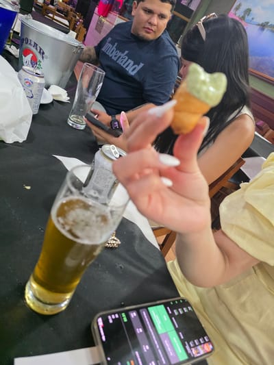 The image shows a close-up view of a table in what appears to be a restaurant or bar, with a portion of a person's hand holding a small food item. On the table, there is a glass of beer with foam, a can of "Polar" beer, an empty glass, and a metal bucket. In the background, a man wearing a navy blue t-shirt with "Timberland" printed on it is visible, along with a woman with long dark hair. A phone screen displaying colorful graphics is also visible in the foreground. The overall setting suggests a casual social gathering.
