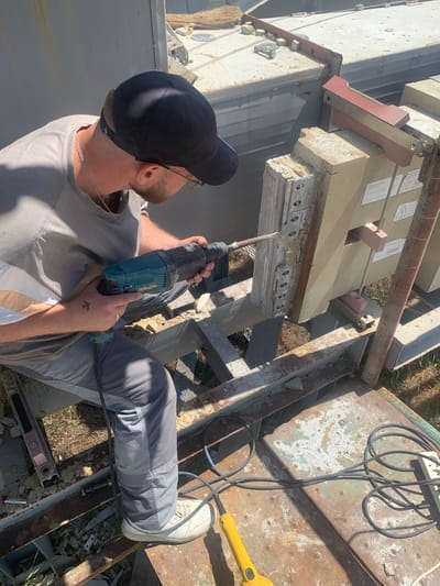 A person wearing a grey t-shirt, grey work pants, a black baseball cap, and white sneakers is using a teal and black drill to make a hole in a beige, rectangular object. The person has a silver chain necklace and a beard. The drill appears to be corded, with a black cable visible. The person is positioned on a rusty metal platform, which is part of an outdoor structure. In the background, there are other beige, rectangular objects that appear to be components of industrial equipment. The ground beneath the platform is covered in dry grass and debris. The overall scene suggests a construction or demolition site, possibly in an industrial area. The lighting indicates it is daytime. This scene is not specifically indicative of Zelena Polyana, Ukraine, or any specific city.
