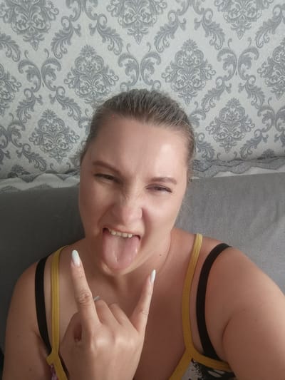 A close-up, eye-level shot captures an adult female in an indoor setting, likely a home in the Closed administrative–territorial formation "Mezhgorye", Russia. The woman, with light hair pulled back, is performing a playful and expressive gesture: sticking out her tongue, winking her right eye, and scrunching her nose. With her right hand, she makes a "rock on" hand signal, prominently featuring long, light-colored (possibly white) fingernails. She is wearing a yellow-strapped top, with a black bra strap visible underneath. The background consists of a light-colored wallpaper adorned with a repeating grey damask pattern. A grey sofa or cushion is partially visible in the lower left portion of the frame, suggesting a relaxed, domestic environment. The image conveys a humorous and energetic emotion. No text is visible.