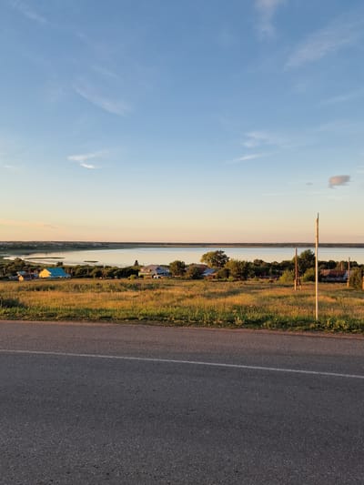 The image depicts a rural landscape under a clear blue sky with wispy white clouds. In the foreground, a grey asphalt road with a white line marking the lane is visible. Beyond the road, a grassy field slopes gently upwards, dotted with wildflowers and tall grasses illuminated by the warm glow of what appears to be sunset. Several houses with pitched roofs are scattered across the field, some with brightly colored roofs. In the middle ground, a tranquil body of water, possibly a lake or river, stretches across the horizon, reflecting the soft light of the sky. A line of trees and vegetation borders the water. A thin, white pole stands vertically on the right side of the image in the grassy field. The overall scene suggests a peaceful, natural environment in a rural area. There are no people or animals visible in the image. The location is not Kashgar City, China; it is a rural landscape.