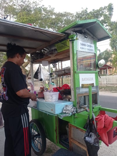 A person with dark, pulled-back hair, wearing a black t-shirt with a graphic design and black pants with red and gray stripes, stands on the right side of a green food cart. The person is looking down and appears to be preparing food. The food cart has a wooden roof and is adorned with signs. One sign reads "TAHU CRISPY BUMBU BALADO" and another reads "BATAGOR". Various food preparation items, containers, and utensils are visible on and within the cart, including a frying pan, bottles of sauce, and plastic tubs. A black plastic bag hangs from the cart, and a red towel is draped over the side. The cart is situated on a paved surface, possibly a street or roadside, with a white picket fence and buildings visible in the background. Trees with green foliage line the area. The scene is set outdoors, with natural lighting suggesting daytime. This scene is located in Desa Talango, Indonesia.