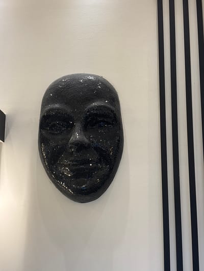 A black, sequined mask is mounted on a white wall. The mask is a human face with visible eyes, nose, and mouth. To the right of the mask, there are black and white vertical stripes on the wall. The background is a plain white wall. The location is identified as a room in the city of Khirbet Al Souk, Jordan.