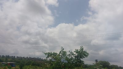The image depicts a cloudy sky above a rural landscape in the city of Nandu, Nigeria. In the foreground, lush green vegetation and trees dominate the lower part of the frame. A small, partially visible structure, possibly a building, is situated on the left side of the image, featuring a blue and red exterior. The middle ground reveals rolling hills covered in scattered trees and vegetation. A few power lines stretch horizontally across the scene. The background is filled with a vast expanse of clouds, varying in density and color from bright white to dark gray, with patches of pale blue sky visible through them. The overall impression is of a natural, somewhat overcast outdoor environment.