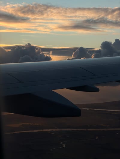 The image captures a view from an airplane window, showcasing a sunset over a rural landscape. The foreground is dominated by the dark silhouette of an airplane wing, angled across the frame. The sky above is a canvas of atmospheric colors, with pastel pinks, oranges, and blues blending with dramatic, dark gray clouds. These clouds are illuminated by the setting sun, creating a sense of depth and texture. Below the clouds, the land appears dark and shadowed, with hints of a winding river or road visible. There are no people or specific landmarks identifiable that would confirm a location in Czerwony Kościół, Poland.