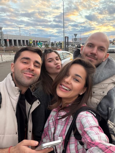Four people are posing for a selfie outside the Yemelyanovo International Airport in Yemelyanovo, Russia. The airport terminal is visible in the background, with the name "АГАФИОН ТЕРМИНАЛ" prominently displayed. Above that, a green and blue logo can be observed. One person has a pink and white plaid shirt. The person taking the photo is wearing a light tan jacket. The sky is cloudy.