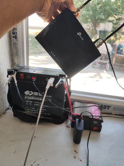 In Sultonobod, Tajikistan, an outdoor scene depicts a setup for powering electronic devices. A black TP-Link router is held by a person's hand, connected to two antennas. This router is positioned near a window, with the outdoors visible through the glass. Below the router, a large black portable power station is placed on a windowsill. This power station, labeled "KM-920" and featuring Chinese text, has jumper cables attached to its terminals, one red and one silver, connecting it to other components. A red indicator light is illuminated on the power station. To the right of the power station, a small black device with a red switch and a USB port is visible, along with a pink power inverter labeled "POWER INVERTER." The overall environment suggests a functional setup with electronic equipment and a power source.