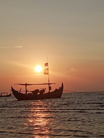 The image displays a sunset over coastal waters in Tritunggal, Indonesia. A prominent, traditional wooden fishing vessel, characterized by a high bow and stern, is silhouetted in the foreground. It features a central mast with a light-colored flag illuminated by the sun, a central canopy structure, and obscured equipment within its hull. A rope or line extends from the stern towards the right. A second, smaller boat is partially visible and silhouetted on the far left. The setting sun, a bright orange disc, is situated low on the horizon, casting a luminous, rippled reflection across the water surface. The sky transitions from a fiery orange and yellow near the sun to a softer blue-grey at the top, with a faint cloud streak present in the upper left quadrant. No specific individuals are discernible within the boats.