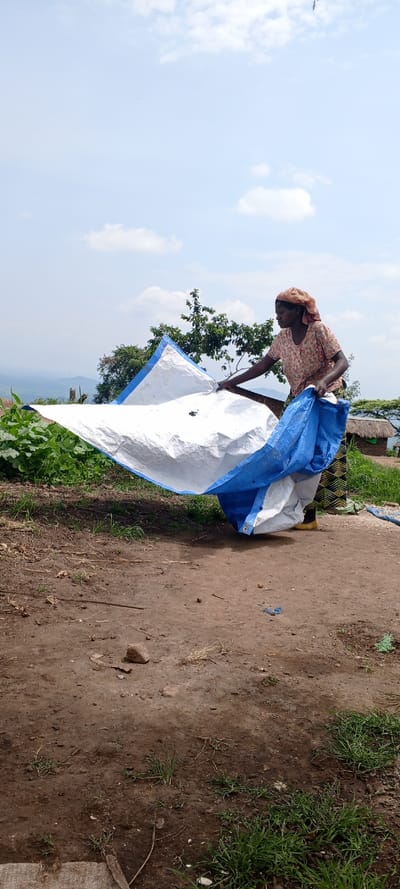 A woman in a patterned blouse and headscarf holds a large, blue and white tarp outdoors. She stands on a dirt patch with sparse grass and rocks. Green vegetation and a tree are visible in the background. In the distance, rolling hills and a cloudy sky suggest a rural or mountainous location. A small, thatched-roof structure is partially visible to the right of the woman.