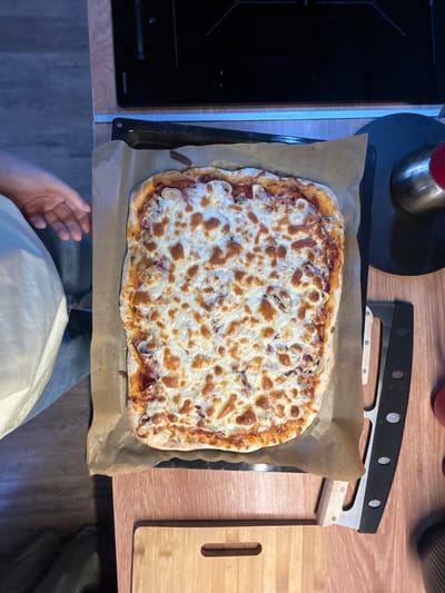 An overhead, close-up shot shows a freshly baked rectangular pizza with melted cheese and visible toppings on a black baking sheet lined with brown parchment paper. The pizza is positioned on a wooden countertop. To the left of the pizza, a person's hand with visible fingers is reaching out, partially obscuring the edge of the pizza. The background includes a portion of a black induction stovetop and other kitchen elements, suggesting the pizza was likely prepared or is about to be served in a kitchen environment. The overall lighting is somewhat dim, with a slight warm cast. The scene appears to be indoors, and given the context provided, it is located in the city of Shepelyovo, Russia.