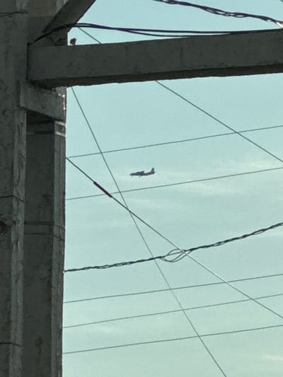 An airplane is seen flying in the sky through a gap in power lines and a concrete structure. The concrete structure appears to be part of a bridge or overpass. The sky is a light blue color, and there are no clouds visible. The airplane is a small, single-engine aircraft. The image was taken in Novomar'evskaya, Russia.