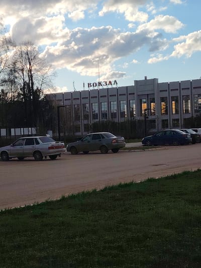 The image presents an outdoor scene in Yanaul, Russia, featuring a large, multi-story building, likely a transport station, as indicated by the prominent red Cyrillic text "ВОКЗАЛ" (VOKZAL, meaning "Station") displayed on its roofline. The building exhibits a robust architectural style, characterized by a grey facade with vertical pillar-like structures and numerous rectangular windows. Reflections of warm, possibly setting or rising sun, cast an orange glow on some of the windows.
In front of the building, several cars are parked on what appears to be a dirt or gravel lot. In the foreground, a light-colored sedan (resembling a Lada model) and a darker, possibly olive-green sedan, are clearly visible. Additional cars are parked further along the lot. To the left of the building and behind the parked cars, there is dense foliage and trees, many appearing bare or sparsely leafed, suggesting either early spring or late autumn. A strip of green grass occupies the bottom portion of the foreground.
The sky above is a bright blue with numerous large, puffy white and grey cumulus clouds, indicating fair weather conditions. The overall lighting suggests either the golden hour of late afternoon or early morning. The scene appears calm and devoid of people, capturing a quiet moment at what is presumably a public transport hub.