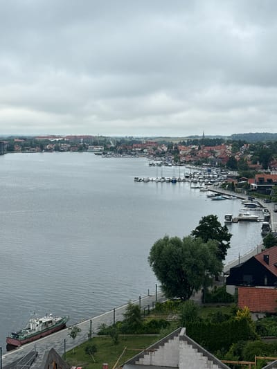 This media file is an aerial view of a harbor in Mikołajki, Poland. The scene depicts a calm body of water filled with numerous boats of various sizes, ranging from small pleasure crafts to larger vessels. The boats are neatly docked along a waterfront lined with buildings and lush green trees. The buildings appear to be a mix of residential and commercial structures, with some featuring red-tiled roofs, characteristic of European architecture. The waterfront promenade is paved, with lampposts and park benches visible, suggesting a public recreational area.
The sky is overcast, indicating a cloudy or perhaps a slightly gloomy day. The water is a muted gray, reflecting the cloudy sky. The overall atmosphere is serene and tranquil, with no visible human activity beyond the boats. A notable detail is a Ferris wheel visible in the distance on the left side of the image, suggesting a town or resort area. The buildings in the background are densely packed, forming a picturesque townscape along the water's edge. The image is taken from a high vantage point, likely from a drone or a tall building, offering a comprehensive view of the harbor and its surroundings. There is no visible text in the image.