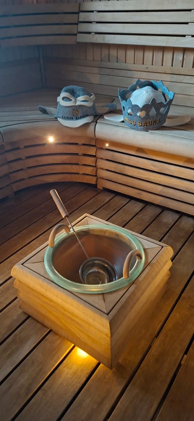 This media file captures an interior view of a sauna in Orimattila, Finland. The scene is dominated by warm-toned wooden planks forming the walls and ceiling, creating a rustic and cozy ambiance. Two sauna hats, crafted from grey felt, are prominently displayed. The upper hat features a decorative grey felt trim with gold lettering that reads "MISS SAUNA". The lower hat has a similar grey felt construction with white accents and lettering that appears to be "SAUNAN KAPTEENI".
To the right, a triangular wooden frame encloses a round, metallic basin with a light green glass rim. A ladle with a wooden handle rests inside the basin, suggesting its intended use for pouring water onto hot stones. The lighting in the sauna is subtle, with small, warm lights embedded in the wooden ceiling, casting a soft glow and creating a relaxing atmosphere. The overall impression is one of traditional Finnish sauna culture, emphasizing relaxation and well-being. The time of day and weather are not discernible from the image.
