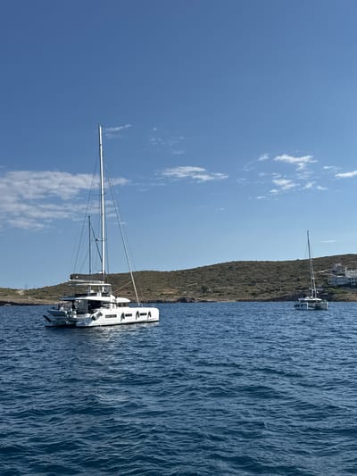The image displays a serene coastal scene under a clear blue sky, consistent with the Aegean Sea within the Lavreotiki Municipal Unit, Greece. Two white sailing catamarans are positioned on the deep blue, gently rippling water. The prominent vessel, a large, modern white catamaran, occupies the mid-ground on the left. It features a tall mast, a furled sail secured on the boom, a hardtop covering the cockpit area, and a grey inflatable dinghy stowed on davits at its stern. Multiple white fenders are visible along its starboard side. Further in the background, to the right, a smaller white sailing catamaran is anchored closer to the shore, also with its mast upright. The horizon is defined by a low, arid-looking landmass, sparsely covered with green and brown scrub vegetation. On the left side of the landmass, a narrow dirt road or track is visible, along with several utility poles. On the right side of the landmass, a cluster of white-painted, multi-story buildings is situated on the hillside. The sky is largely clear, with scattered patches of small, white cumulus clouds. Bright sunlight illuminates the scene, indicating good weather conditions. No people are discernible on either vessel or on the land.