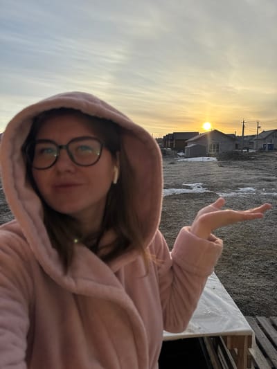 A selfie-style photograph shows a woman with glasses and a pink hooded robe. She is wearing a wireless earbud in her left ear and making a hand gesture. The background features a landscape including houses, utility poles, and a sky with a setting or rising sun. The ground has a patchy appearance, possibly snow or frost. The location is Nestiukovo, Russia, based on the prompt.
