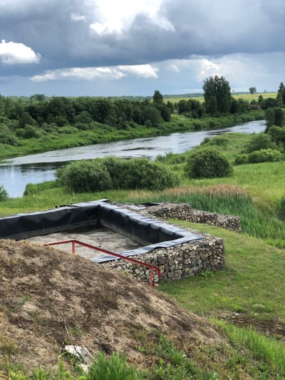 The image depicts an outdoor scene in Nadezhda, Russia, featuring a river flanked by lush green vegetation and a dramatic, cloudy sky. In the foreground, a partially constructed stone-lined pit with a black liner and red metal supports is visible. The pit is situated on a grassy slope, with dry soil and sparse vegetation at the very front. Adjacent to the pit are more stone-lined structures, possibly gabions, and tall reeds. The river flows through the middle ground, bordered by dense trees and bushes on both sides. The background consists of a field and more trees, extending towards the horizon. The overall impression is a natural landscape under a moody sky, with evidence of construction in the foreground. No people are present in the image.