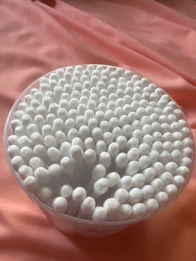 The image shows a clear plastic container filled with cotton swabs, arranged in a dense, somewhat circular pattern. The swabs are white, with rounded cotton tips. The container is placed on a wrinkled, pale pink fabric that appears to be silk or satin, casting soft shadows. The lighting is soft and diffused, highlighting the texture of both the cotton swabs and the fabric. The background is entirely composed of this pink fabric, with some areas appearing darker due to folds and shadows. There are no people or animals visible in the image. The image does not provide any information or context that would allow for its location to be determined in the city of Pskov, Russia.