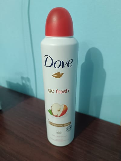 A Dove Go Fresh deodorant spray is positioned upright on a dark wood surface. The deodorant can is white with a red cap and features the Dove logo, the text "go fresh," an illustration of a sliced apple, and information about "moisturising cream" and "48h." The background is a light blue wall with a shadow of the deodorant can cast on it. The image was taken in Vabkent, Uzbekistan.