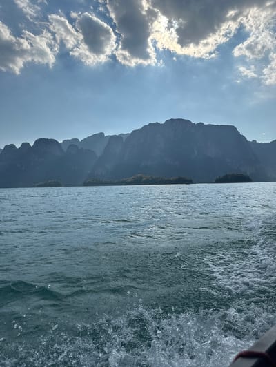 The image depicts a waterscape in Ban Chiao Lan, Thailand. On the left, a dramatic mountain range rises against a sky with scattered clouds. The right side of the image is filled with water, disturbed by the movement of a boat, evident in the spray and ripples.
