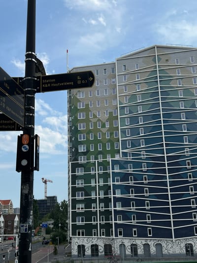 The image shows a street scene in Zaandam, Netherlands, dominated by a modern building with a geometric facade of green and blue panels and white diagonal supports. The building is tall and appears to be residential or office space. In the foreground, a black lamppost with multiple directional signs is prominent. These signs provide directions to various locations, including "Station K&R Houtveldweg," "Stadhuis Busstation," "Taxi K&R Noordschebos," and "theater". One sign indicates "KOR 0.1" with an arrow pointing upwards.
The sky is partly cloudy, suggesting it is daytime, possibly late morning or early afternoon. A construction crane is visible in the distance behind the building, indicating ongoing development in the area. The overall scene is urban, with elements of modern architecture and infrastructure. No people are clearly visible in the frame, though the presence of the street signs and building suggests a public urban space. The image is oriented vertically, as if taken from a low angle looking upwards.