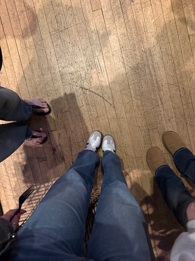 A floor shot shows three pairs of legs and feet. At the top, legs clad in jeans and tan boots angle toward the right. A similar pair of legs in jeans and sneakers is positioned towards the center of the frame. The bottom pair of legs, also in jeans, wears black flip-flops revealing pale toes, complete the trio. A hand holding a black fork peeks in from the right edge, indicating someone is at a table. The floor is made of light wood with visible wear and tear.