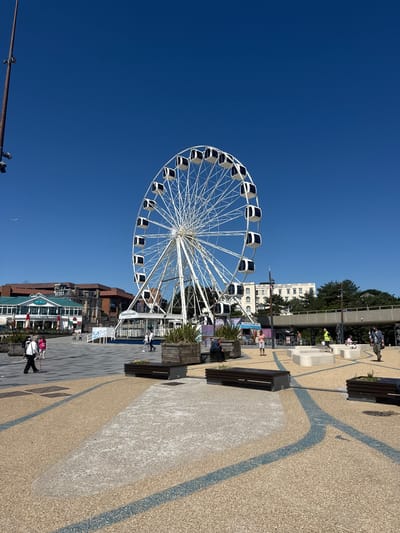This is a medium-shot, eye-level, outdoor, vertical photograph taken on a bright, sunny day in Bournemouth, United Kingdom. The image features a large white Ferris wheel prominently placed in the center of the frame, against a clear, deep blue sky. The Ferris wheel has many cabins, each with dark windows. Below the Ferris wheel, there is a plaza with a patterned ground surface composed of light tan and grey sections, interspersed with blue lines. Several dark wooden benches are positioned around the plaza. In the background, there are buildings of varying architectural styles, including a green-roofed structure with the signage "The Rock," and a white, multi-story building that appears to be a hotel. Trees are visible behind the buildings. Several people are scattered throughout the plaza, some walking, some sitting on benches, and others standing. One person in the mid-ground is wearing a high-visibility yellow vest. The overall scene depicts a public space in an urban coastal area.