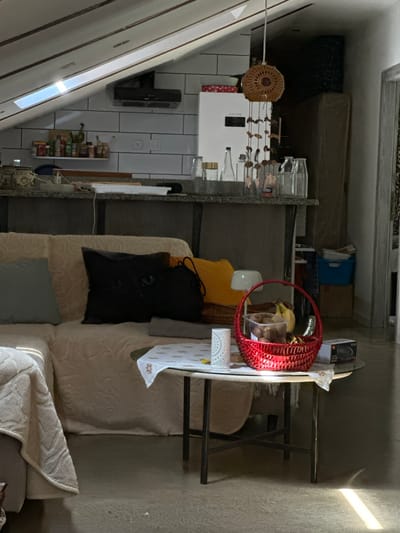 This media file depicts the interior of a lived-in room, likely a combined living and kitchen area, in Bartula, Montenegro. The scene is brightly lit by natural sunlight, with strong rays and shadows cast on the floor and furniture, indicating a sunny late morning or afternoon.
In the foreground, a light-colored sofa or couch is visible, adorned with cushions in yellow, black, and light blue, and covered with a beige patterned blanket. Adjacent to the sofa, a dark metal table holds a white tablecloth. On the table, a vibrant red woven basket contains bananas and other fruits, alongside a clear container holding cookies or pastries. A white table lamp and a black box for a JBL speaker, with visible text "JBL" and "10.360," are also present.
Beyond this seating area, the room features a section with white subway-style wall tiles. A wall-mounted spice rack, filled with numerous jars, is prominent, as is a dark appliance, possibly an oven or microwave, mounted higher on the wall. A countertop below displays various glass bottles and a clear glass pitcher. From the ceiling, a round, woven decorative piece and a wind chime-like mobile hang.
The background reveals stacked items, possibly mattresses or other storage, against a wall. The room features a distinctive sloped ceiling with white paneling and visible beams, typical of an attic or top-floor dwelling, with a skylight letting in light. The floor is light-colored. No people are visible, and no specific activity is in progress, portraying a static, homely interior.