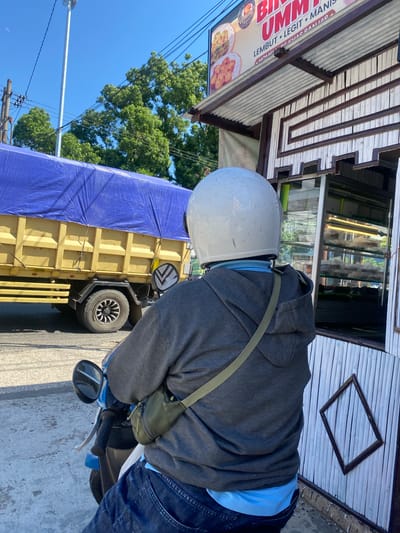 A person wearing a white helmet and a grey hoodie sits on a blue and white motorcycle, facing away from the camera. They are positioned next to a small, open-air food stall constructed with bamboo siding and a corrugated metal roof. A sign above the stall reads "BIM UMUM LEMBUT LEGIT MANIS." In the background, a large yellow truck covered with a blue tarp is parked on the side of a street. Trees and utility poles are visible behind the truck. The scene appears to be outdoors, with a clear blue sky overhead. The location is identified as Sungai Paring, Indonesia.
