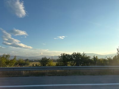 The image depicts a view from a roadside, likely a highway, looking towards a landscape. In the foreground, a concrete road surface with a white line is visible, followed by a metal guardrail. Beyond the guardrail, there is a grassy area with some dry vegetation, and then a line of trees and bushes. In the middle ground, a field stretches out, and further in the distance, there are more trees, hills, and mountains under a clear blue sky with scattered clouds. The sun appears to be setting or rising, casting a warm light on the scene. The overall impression is one of a rural or semi-rural environment. There are no people or specific architectural elements that would definitively place this scene in Dzveli Kanda, Georgia.