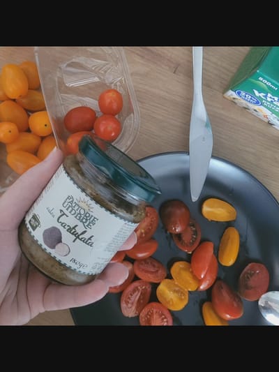 In Bielawska Wieś, Poland, an overhead shot displays a table with various food items. A clear plastic container holds a collection of cherry tomatoes, some yellow and some red. Beside it, a jar of "Fattorie Uidbre Salsa Tartufata" is held in a person's hand. The jar's label is visible, featuring an illustration of truffles and text indicating the product is 180g. A silver butter knife rests on a dark grey plate filled with halved cherry tomatoes, both red and yellow varieties. In the background, a portion of a green box with white and blue text that reads "KM 500ml" is visible. The overall scene is a preparation of a meal or snack, likely involving fresh tomatoes and a truffle-based salsa.