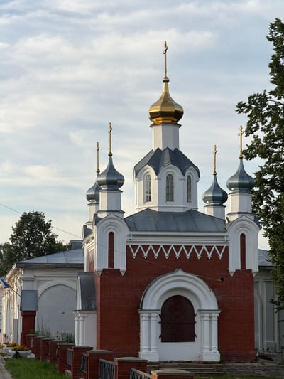 This is an exterior, eye-level, medium shot of a red brick church with a white facade and several onion domes, located in Cherdyn, Russia. The church has a central, larger dome that is gold, and four smaller domes that are silver. The domes are topped with golden crosses. The church has arched windows and decorative white trim along the roofline. The building is surrounded by some greenery, with trees visible on the right side of the image and some potted flowers along a low fence in the foreground on the left. The sky is overcast with soft, white clouds. There are no people or animals visible in the image.