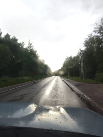 The image captures a view from inside a vehicle driving on a wet, asphalt road. The road appears to be bordered by dense green trees on both sides, with power lines running alongside the trees. The sky is overcast and bright, suggesting either dawn or dusk, or a heavily clouded day. There are visible water droplets on the windshield, indicating that it might be raining or has recently rained, contributing to the wetness of the road and the reflections on its surface. The light from the sky reflects strongly on the wet road, creating a bright streak down the center. The hood of the vehicle is visible in the foreground, reflecting the sky and the surrounding trees. The overall impression is that of a journey through a rural or semi-rural landscape during possibly inclement weather. The location context provided is Khodaevo, Russia. There is no visible text in the image.