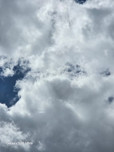 This is a photograph of a cloudy sky. The clouds are a mix of white and grey, with some areas of darker grey suggesting thicker cloud cover. Patches of deep blue sky are visible through breaks in the clouds. The lighting suggests it is daytime, possibly late afternoon or early evening given the diffused light. The scene is a natural outdoor setting, with the sky filling the entire frame. There are no people or man-made objects visible, focusing solely on the atmospheric conditions. The overall mood is somewhat dramatic due to the contrasting light and dark clouds. Text is visible in the lower right corner of the image, oriented vertically, which reads "Galaxy S26 Ultra".