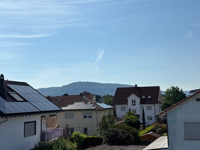 This media file depicts a bright, sunny daytime scene overlooking a residential area in Wörth am Main, Germany. The foreground features the angled rooftops of several houses, with one prominently displaying a large array of dark solar panels. Another house in the middle ground also has solar panels installed on its roof. The houses are of varying architectural styles, predominantly featuring red-tiled roofs and white or light-colored walls. Lush green trees and bushes surround the houses, indicating a well-maintained residential environment.

In the background, a large, tree-covered hill or mountain range rises against a clear blue sky. The sky itself shows some wispy clouds, and a faint contrail from an airplane is visible, suggesting travel. The overall atmosphere is serene and peaceful, typical of a pleasant day in a suburban or semi-rural setting. There are no people visible in the image. The scene is captured from an elevated perspective, looking down upon the rooftops and across the landscape. The time of day is clearly daytime due to the bright sunlight and clear visibility. The presence of solar panels suggests an environmentally conscious community. No text is visible in the image.