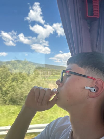 The image captures a close-up profile of a young person, likely male, seated next to a window, looking out to the left. The person has short dark hair, is wearing black-framed glasses with red accents on the temples, and a white t-shirt. A white earbud (resembling Apple AirPods Pro) is visible in their right ear. Their right hand is raised, with fingers touching their chin, suggesting a moment of thought or contemplation.
Through the window, a bright, clear blue sky with scattered white cumulus clouds dominates the upper half of the outdoor scene. Below the sky, a verdant landscape stretches out, filled with green foliage, possibly fields or hills, hinting at a rural or natural environment. The indoor setting suggests the person is inside a vehicle, possibly a bus or train, indicated by a deep purple curtain and a red plastic handle (part of a window or shade mechanism) visible at the top of the frame.
The scene depicts a sunny day with good weather. The overall impression is one of quiet observation or travel. Based on the provided context, the location is Nea Karvali, Greece. No discernible text is visible in the image.